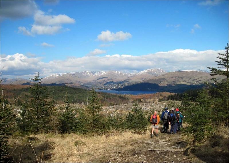 A group of hikers on a mountain path with a scenic view of forests, a lake, and snow-capped peaks in the distance.