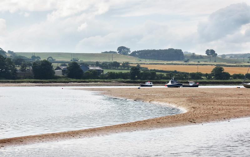 Alnmouth | Boats on a calm river with a sandy shore and rolling green hills in the background.