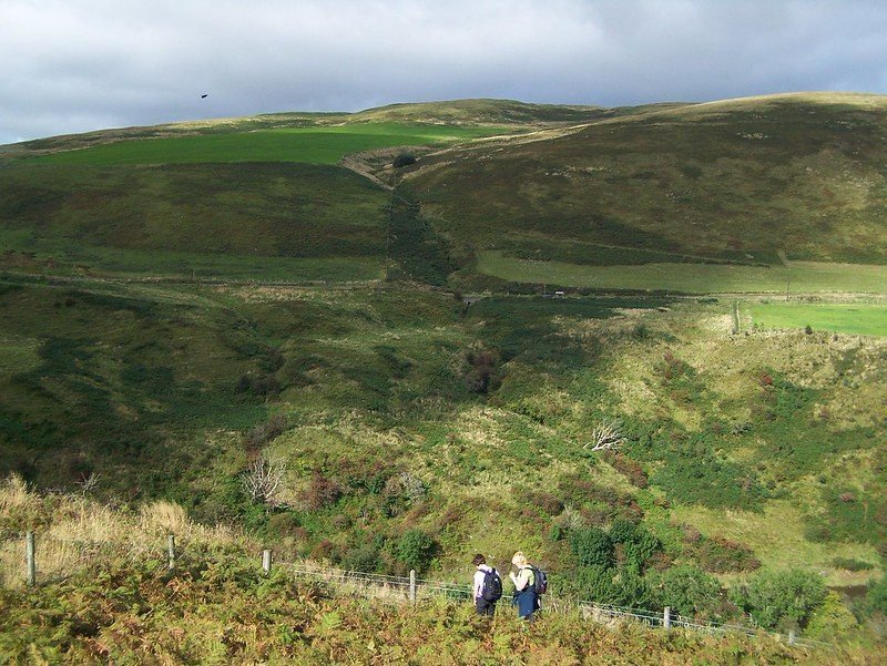 Two hikers walking down a grassy hill, surrounded by green, rolling hills under a cloudy sky.