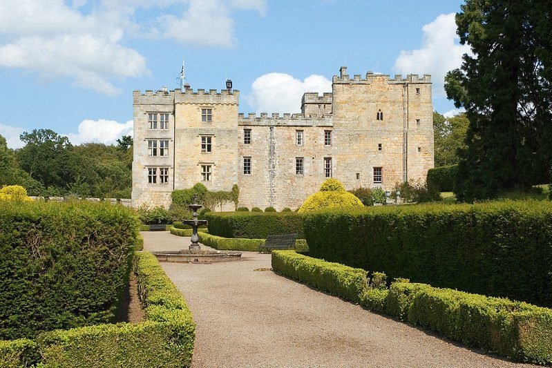 A historic stone castle with crenellated towers is surrounded by manicured gardens and a gravel pathway leading to a central fountain.