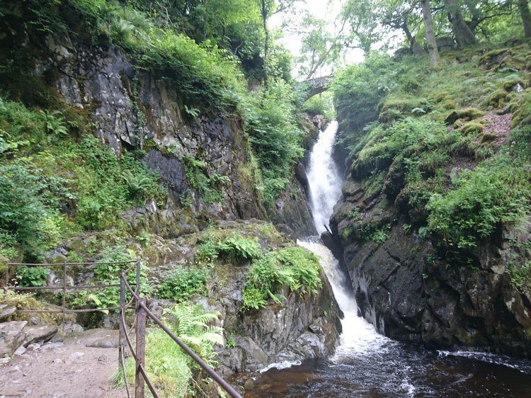 Helvellyn | Guardian of Glenridding and Patterdale 3 Waterfall flowing between steep rocky cliffs covered with green vegetation and moss, with a metal railing and pathway on the left side.