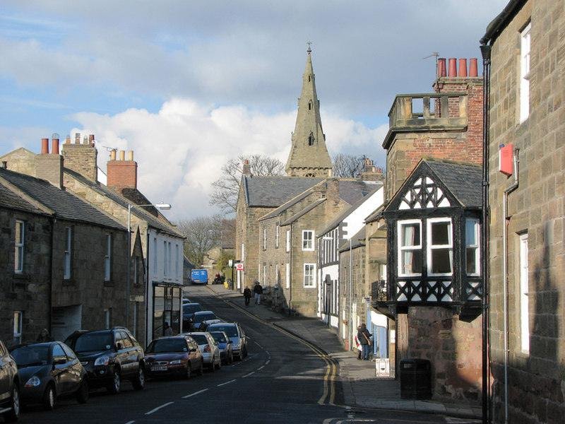 Street view of a quaint village with stone buildings, parked cars, and a church steeple in the background.