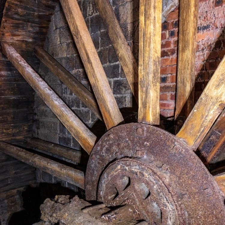 Close-up of a large rusted metal and wooden water wheel with a brick and stone wall background, showing weathered wooden spokes and worn metal components.