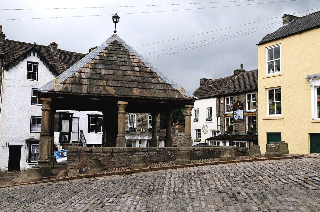 Stone market cross with a shingled roof supported by pillars on a cobbled street, surrounded by traditional white and yellow buildings under a cloudy sky.