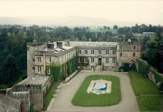 A large historic stone mansion with a central courtyard and landscaped garden, surrounded by lush green countryside and distant hills.