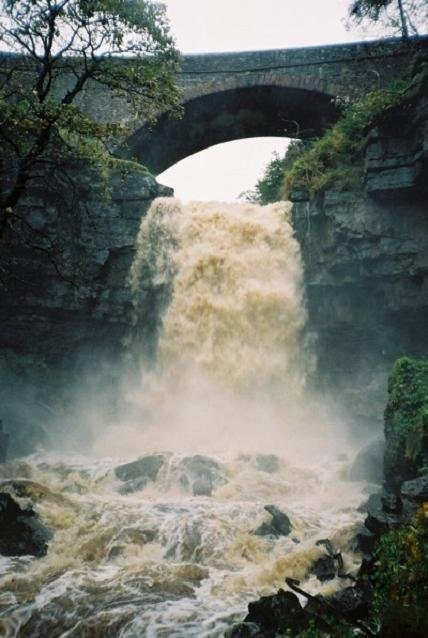 Powerful waterfall cascading over rocky cliffs beneath a stone arch bridge surrounded by trees and greenery
