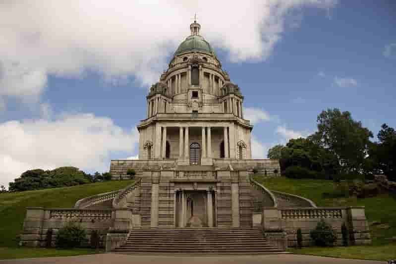 A grand neoclassical building with a large dome and ornate stonework, surrounded by green lawns and trees under a partly cloudy sky.