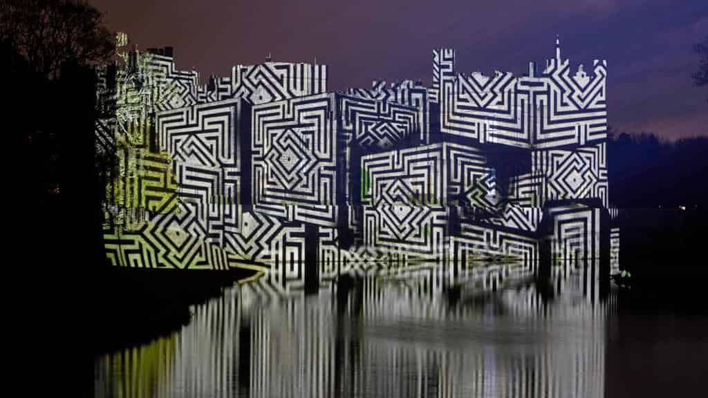 A waterfront building at night covered in bold black-and-white maze-like geometric projection patterns, the illuminated façade reflected in the calm water with dark tree silhouettes to the left and a purple night sky above.