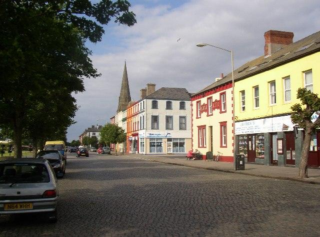 Criffel Street, Silloth, A street with colourful buildings and small shops, featuring a church spire in the background, parked cars along the side, and a tree-lined sidewalk.