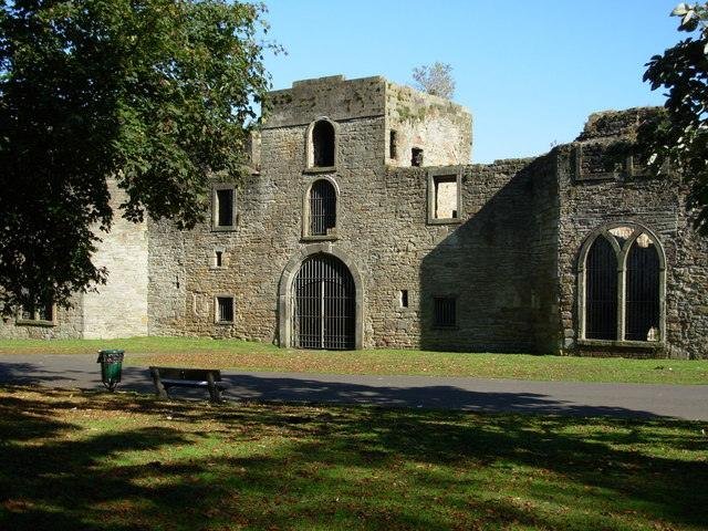 Workington | Ruins of an old stone building with barred windows and arched doorways, surrounded by trees and a grassy area with a bench and bin.