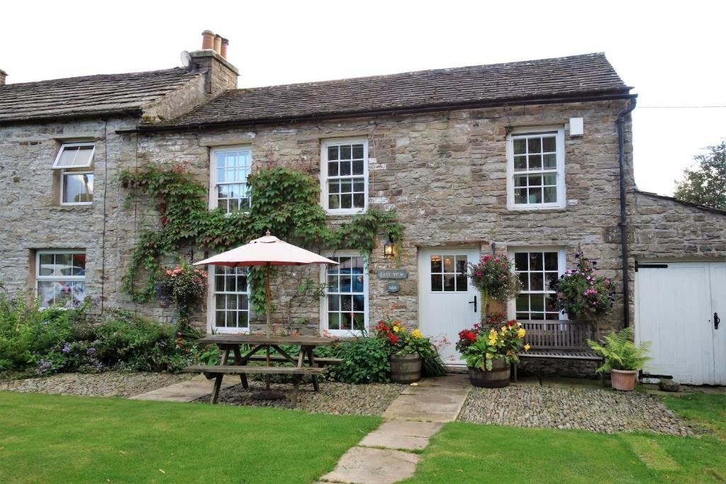 Traditional stone cottage with white-framed windows and door, climbing green ivy, hanging flower baskets, a wooden picnic table with a large parasol, and a wooden bench, set in a garden with green grass and a stone pathway leading to the house.