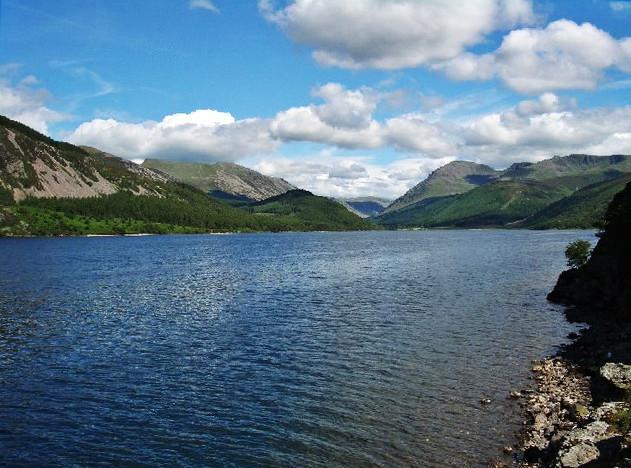 A serene lake surrounded by green hills and mountains under a partly cloudy sky.