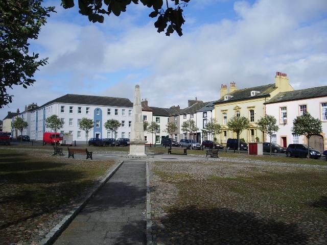 Fleming Square, Maryport | Town square with a tall stone monument in the centre, surrounded by colourful buildings, parked cars, benches, and small trees.