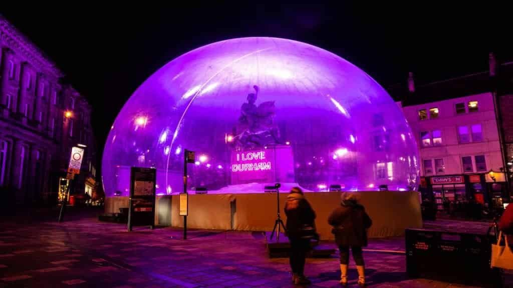 Large translucent inflatable dome glowing purple in a nighttime city square, enclosing a statue and a neon sign that reads I LOVE DURHAM, with two bundled-up figures in the foreground and surrounding buildings lit by purple lights