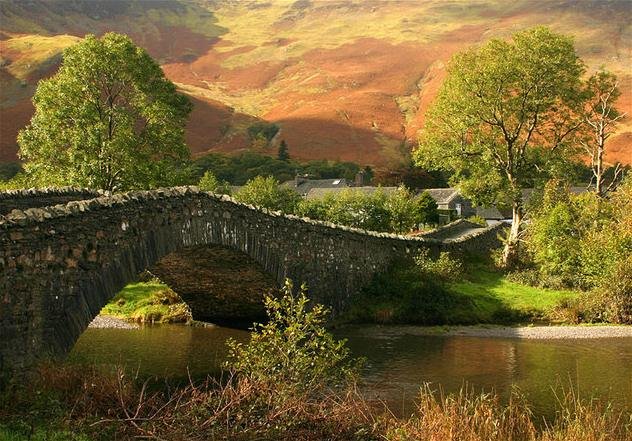 Stone bridge over a river in an autumnal landscape with trees and hill in the background.