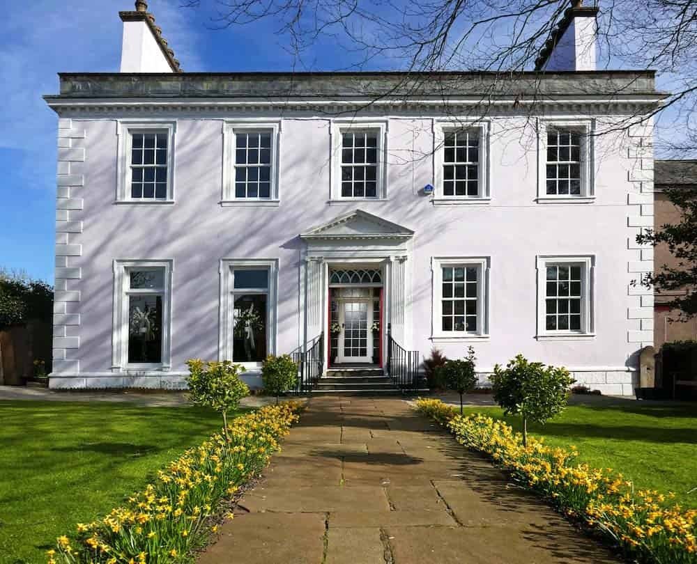 Pale lilac Georgian house, the Helena Thompson Museum, with white trim and a red door, seen on a sunny day with a stone path flanked by yellow daffodils leading to the entrance.