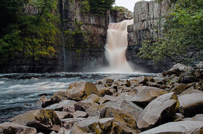 Waterfall cascading between rocky cliffs into a river surrounded by large rocks and greenery