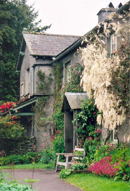 Stone cottage with lush greenery, flowers, and a bench in front.