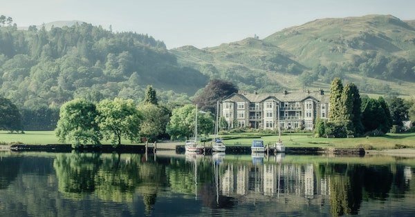 Affiliate Disclosure 1 Large stone building surrounded by trees on a grassy area in front of hills, with several boats docked on a calm lake reflecting the scene