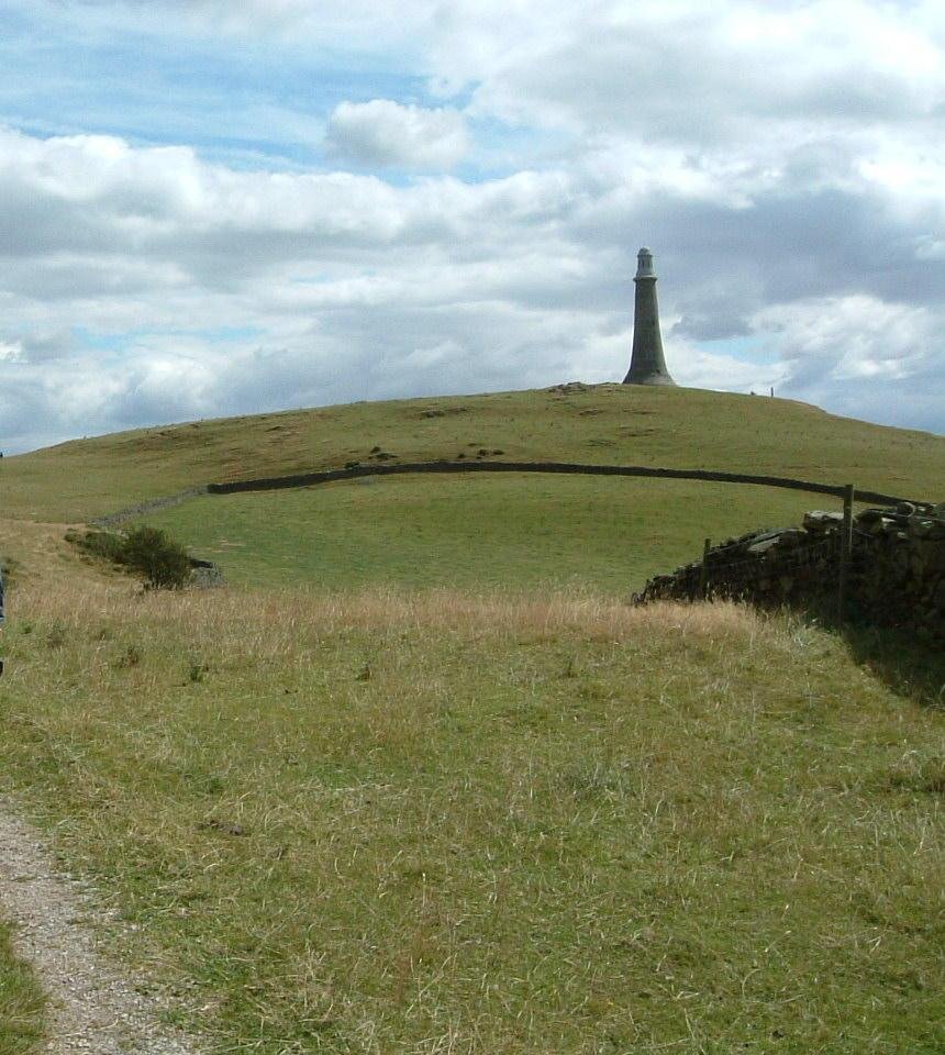 A stone monument atop a grassy hill under a cloudy sky, with a winding path and dry stone walls in the foreground.