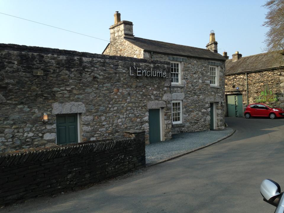 Stone building with the sign "L'Enclume" above the door, a red car parked beside a garage with a sign for "Rall Tech", and a small gated area in front.