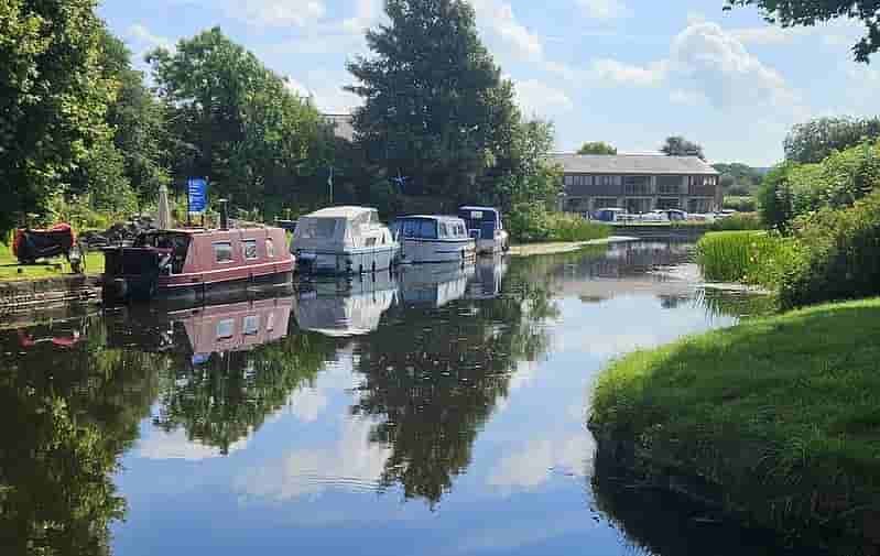 Canal with moored boats surrounded by greenery and a building in the background.