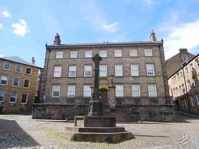 A historic stone building with multiple windows, featuring a cross monument in front and surrounding cobblestone pavement under a blue sky.