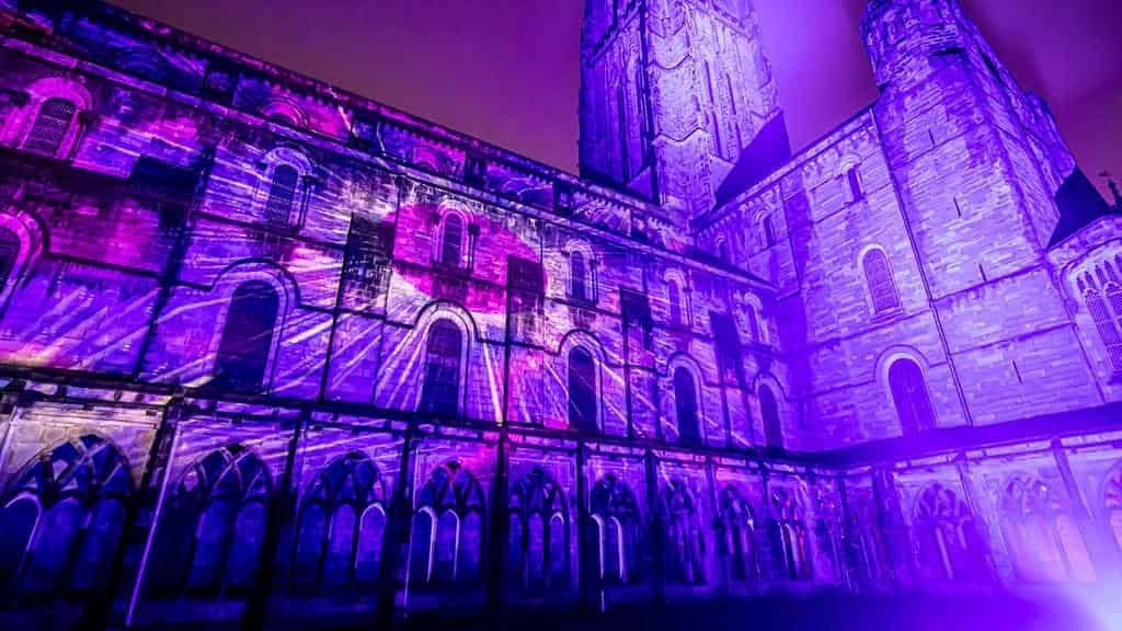 Gothic cathedral exterior and cloister lit by vivid purple and magenta light projections sweeping across its stone arches, windows and tower at night.