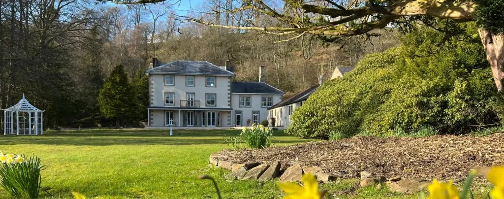 Large two-story house with a white gazebo on the left and a garden with blooming daffodils and green bushes in the foreground under a clear blue sky