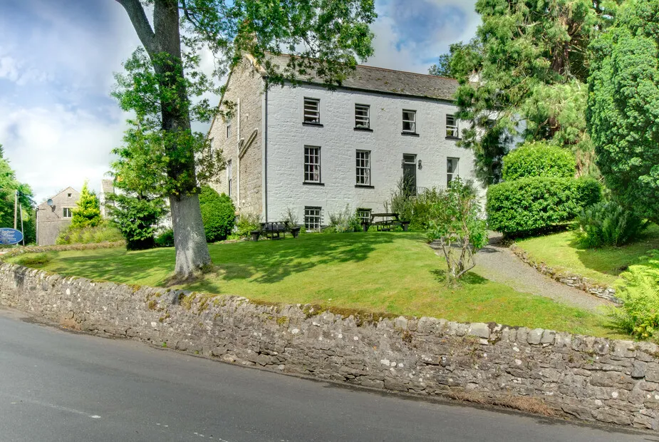 White two-storey stone house with black-framed windows and a lawn in front surrounded by tall trees and bushes, bordered by a low stone wall next to a road