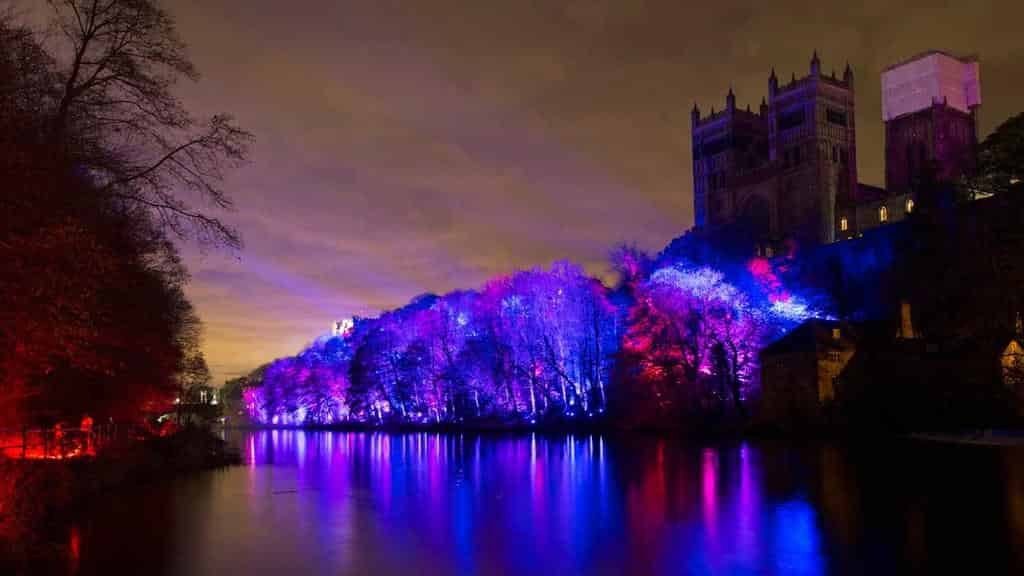 Durham Lumiere 2025: Night scene of a river with trees lit in purple and pink lights reflecting on the water, and a large stone cathedral-like castle perched on a hill above the illuminated bank.