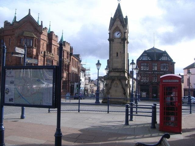 Town square with a clock tower, red telephone box, a map board, and historic buildings in the background.