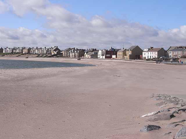 Newbiggin-by-the-Sea, Sandy beach with calm water, a row of houses in the background, and partly cloudy sky.