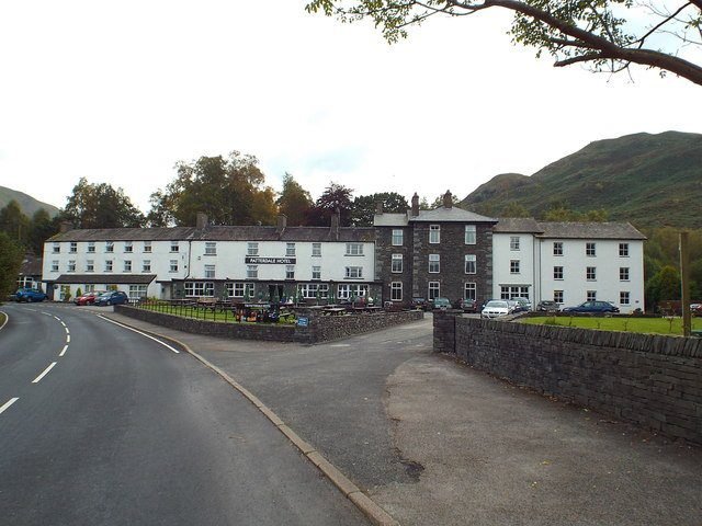 Helvellyn | Guardian of Glenridding and Patterdale 5 Exterior view of a large multi-story hotel building with white and dark stone walls, surrounded by trees and hills under an overcast sky, with a road curving past the front and several parked cars