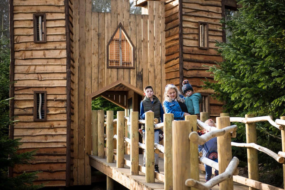 Two children and a woman walking on a wooden bridge leading to a rustic wooden cabin surrounded by trees