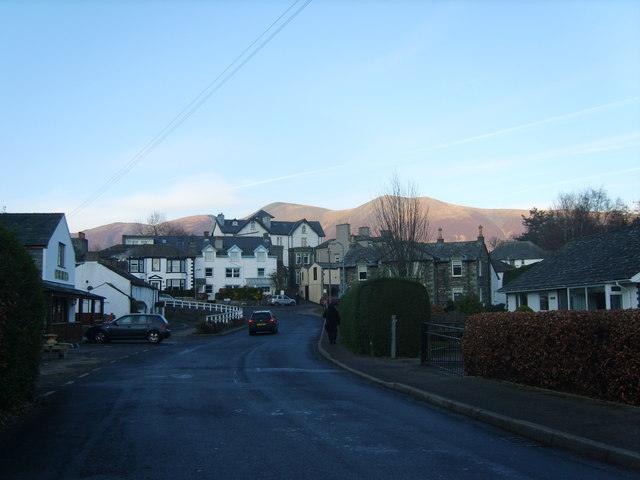 Portinscale, Cumbria | Road leading through a quaint village with traditional houses and hills in the background.