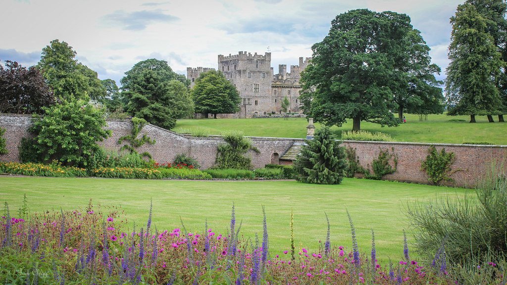 Historic stone castle in the distance behind a brick wall with lush green trees and well-maintained gardens featuring vibrant purple and pink flowers in the foreground.
