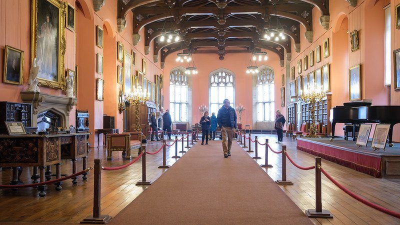 Spacious historic gallery with pink walls, wooden floors, and a high ornate ceiling, lined with framed paintings, antique furniture, chandeliers, and a grand piano, with several people walking and observing the exhibits.