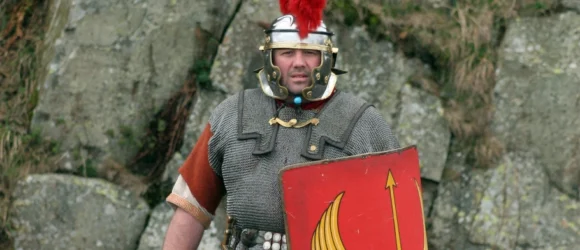 A man dressed in Roman legionary armour holding a red shield with a yellow design.