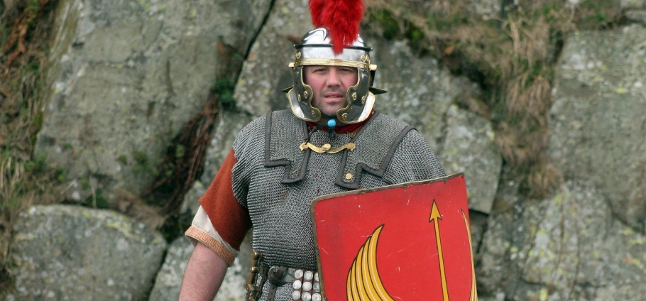 A man dressed in Roman legionary armour holding a red shield with a yellow design.