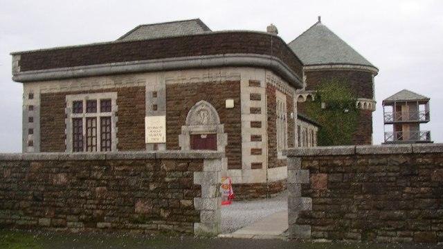 Stone building with rectangular and arched windows, surrounded by a brick wall, featuring a round turreted structure slightly visible behind it.