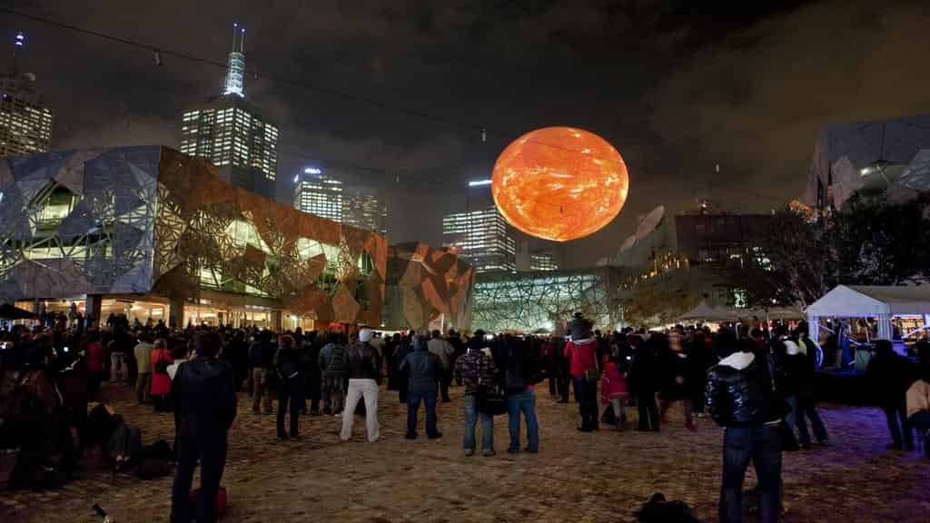 Large crowd gathered at night in a city square with reflective angular buildings and skyscrapers, watching a huge illuminated orange orb suspended above the plaza.