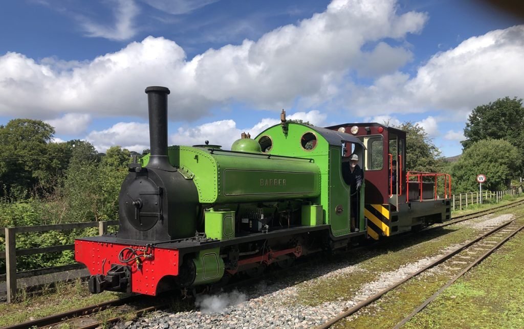 Green and black steam locomotive named Barber on railway tracks with a person in the driver's cabin, surrounded by greenery under a partly cloudy blue sky.