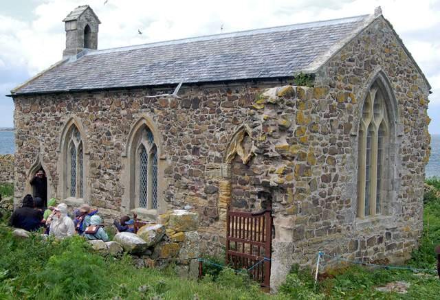 Stone chapel with a small group of people gathered outside, surrounded by lush greenery and a view of the sea in the background.