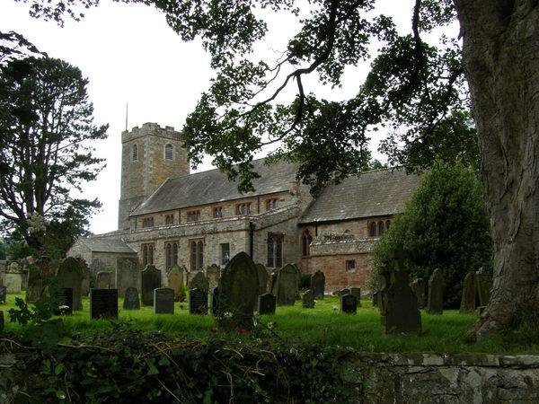 A stone church with a large tower surrounded by a graveyard, framed by trees.