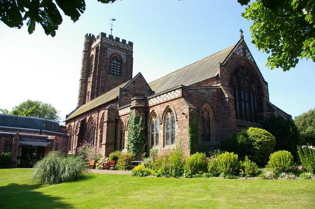 A red sandstone church with a square tower and stained glass windows, surrounded by a well-kept garden.