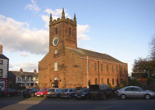 St Mary's Church, Wigton | Stone church with a clock tower and arched windows, surrounded by parked cars under a blue sky.