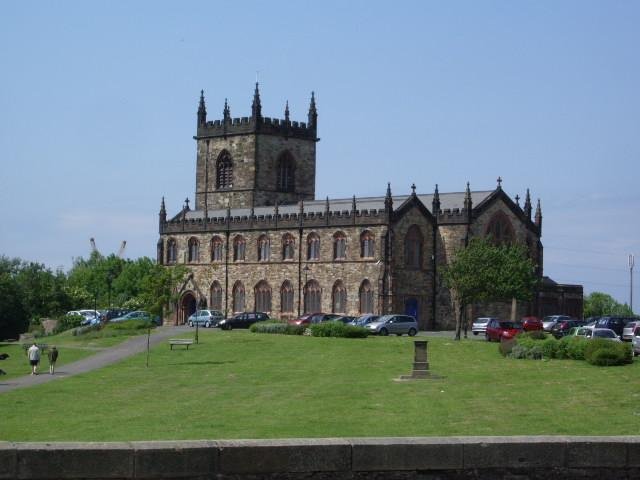 Stone church with a square tower and crenellations, set in a grassy area with parked cars and walking paths.