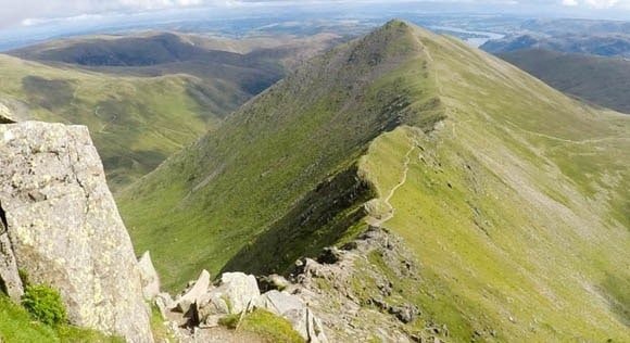 Helvellyn | Guardian of Glenridding and Patterdale 2 A grassy mountain ridge with a rocky outcrop in the foreground and a winding trail leading to the summit under a partly cloudy sky
