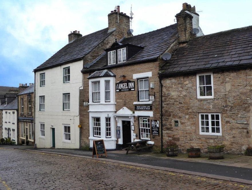Cobblestone street with a row of old stone buildings including the Angel Inn pub with signs and a picnic table outside on a cloudy day